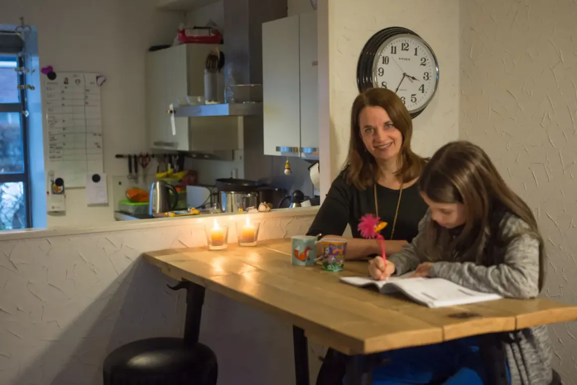 Moeder helpt dochter met huiswerk, terwijl kaarsen branden. Op de muur hangt een klok en een whiteboard met een verhoudingstabel procenten.