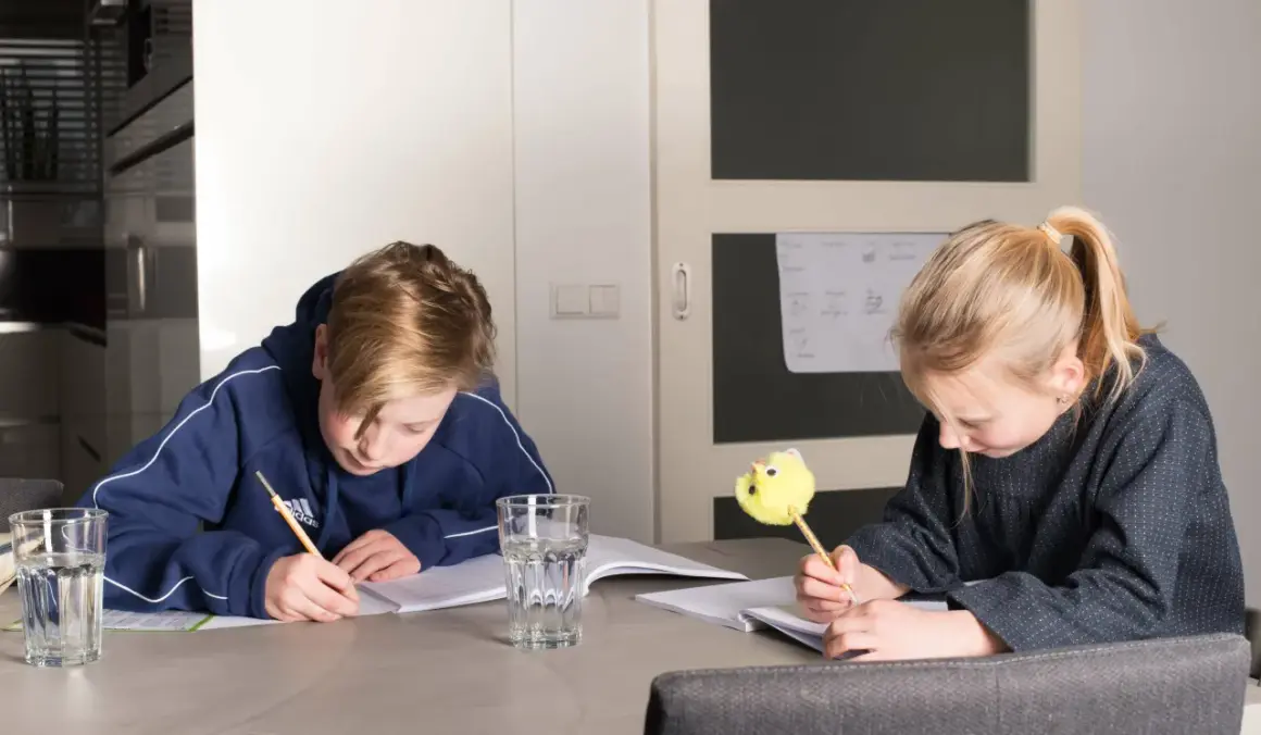 Twee kinderen maken huiswerk aan tafel. De jongen schrijft met een potlood, de meid met een leuke gele pen. Ze oefenen met delen met kommagetallen.