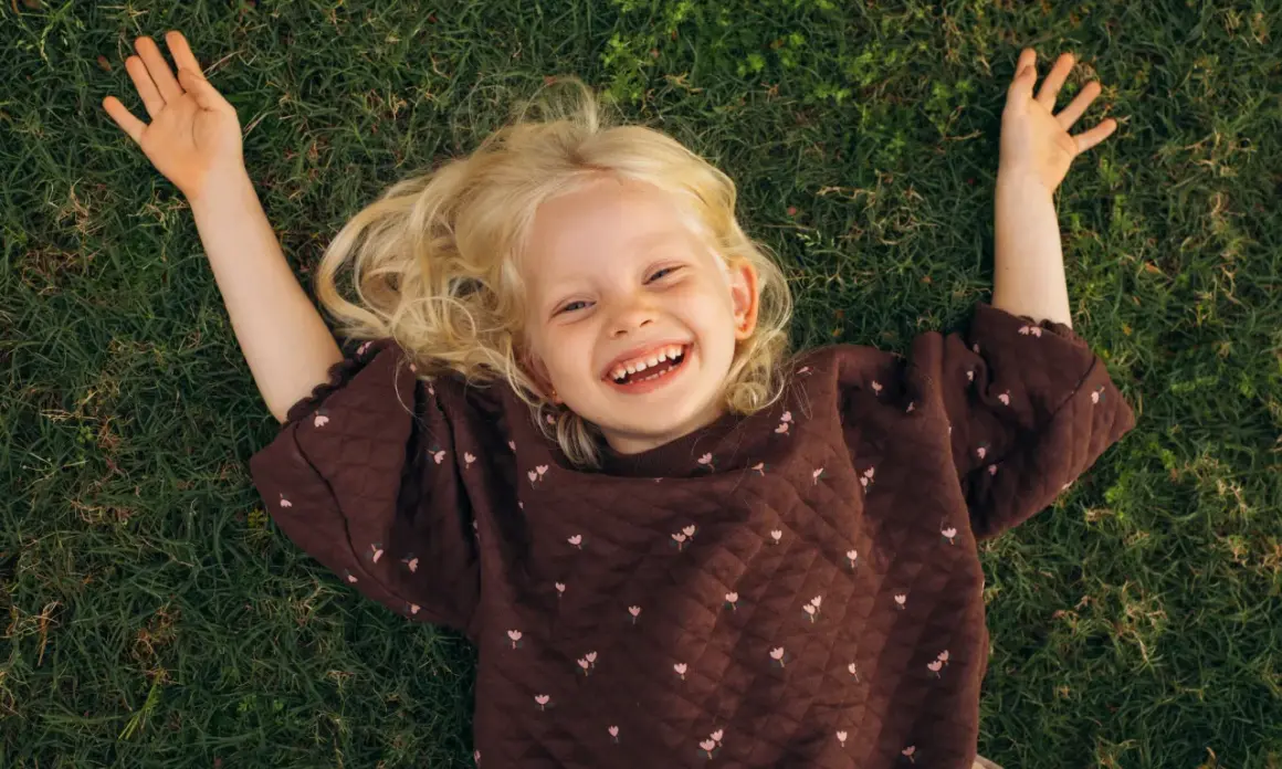 Lachend meisje met blonde krullen ligt op gras. Ze geniet van de rust, een moment van kalmte bij onderprikkeling ADHD.