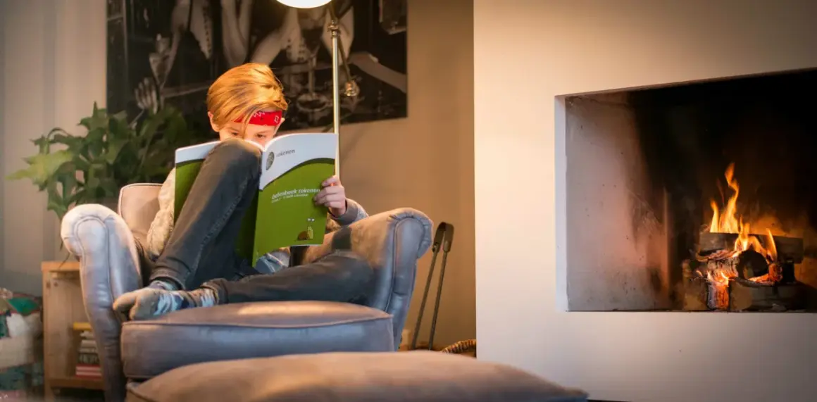 Kind met rood bandje leest een oefenboek rekenen bij de haard.