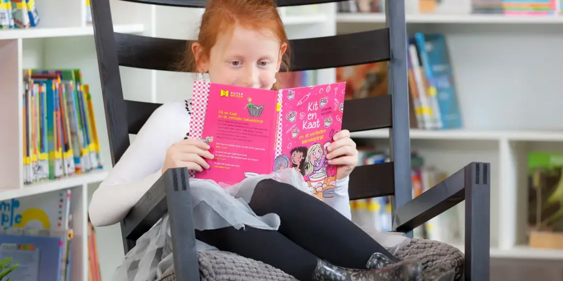 Een meisje met rood haar oefent met lezen in een comfortabele stoel, omringd door boeken.