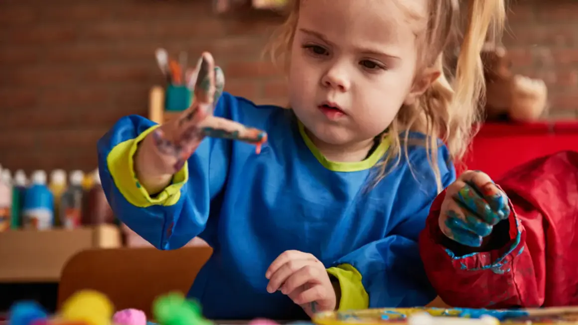 Een peuter met verf aan haar handen, die geconcentreerd aan het schilderen is. Dit is een mooi voorbeeld van cognitieve ontwikkeling peuter door creatief spel.