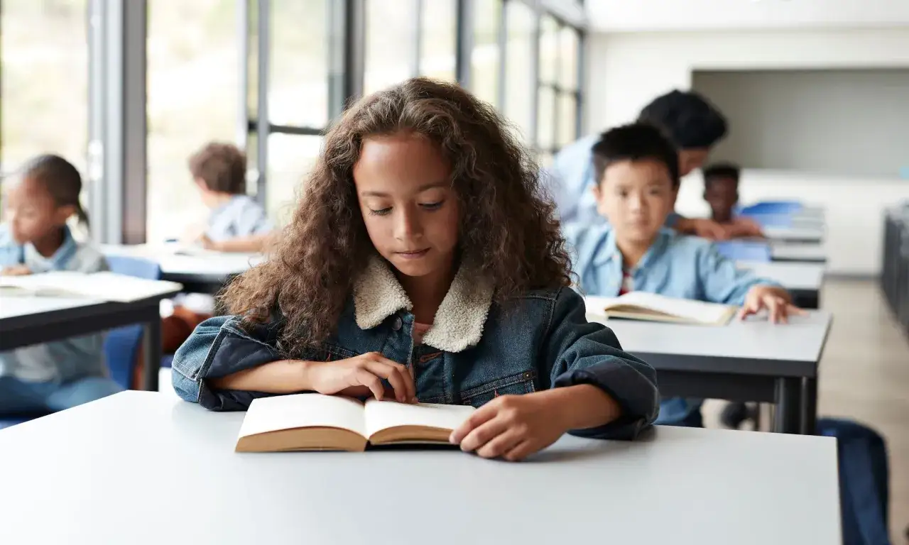 Een meisje met krullend haar leest geconcentreerd in een boek. Ze lijkt diep in gedachten, een mogelijke uiting van symptomen hoogbegaafdheid.