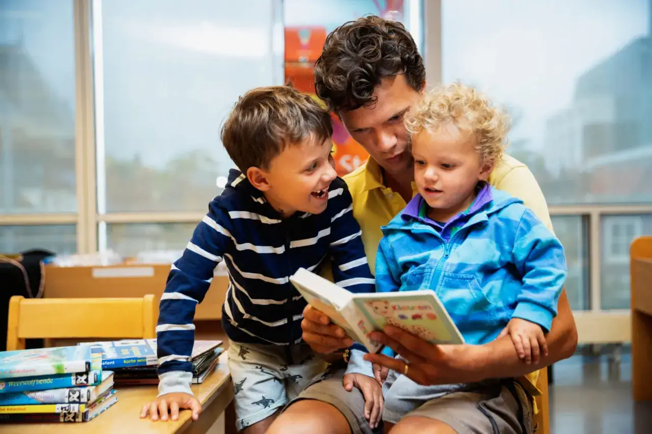 Vader leest met zijn kinderen een boek voor. De oudste zoon lacht, de jongste luistert aandachtig. Een vrolijk moment, net als de leeftijdsgrens basisschool 1 januari.