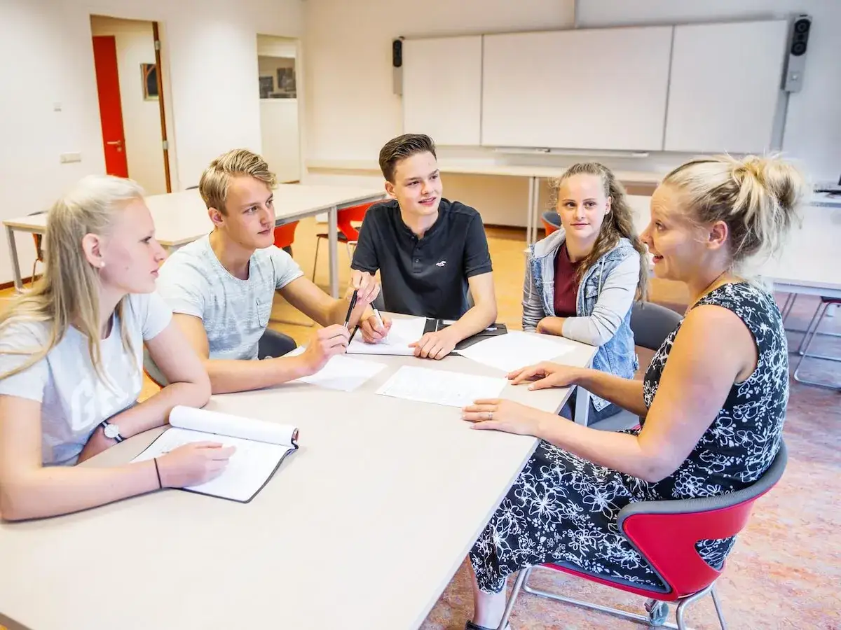 Een docent is in gesprek met de leerling aan een tafel. Ze kijken elkaar aan en er liggen papieren.