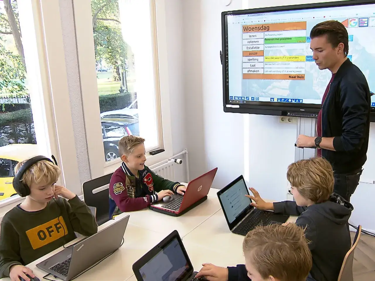 Kinderen leren op laptops in een klaslokaal van een particuliere basisschool. Een leraar wijst naar een digibord met het lesrooster.
