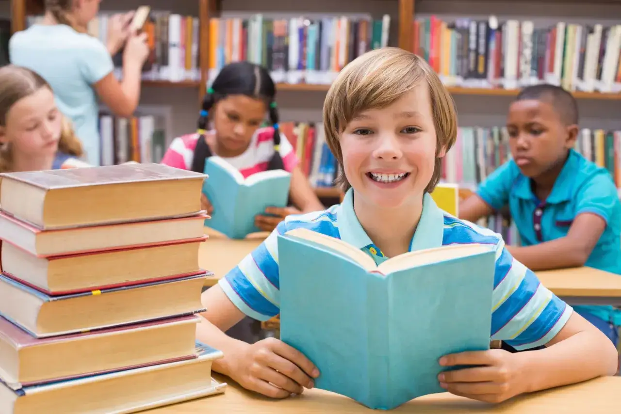 Een lachende jongen leest een boek in de bibliotheek, omringd door stapels avi plus boeken en andere kinderen die ook lezen.