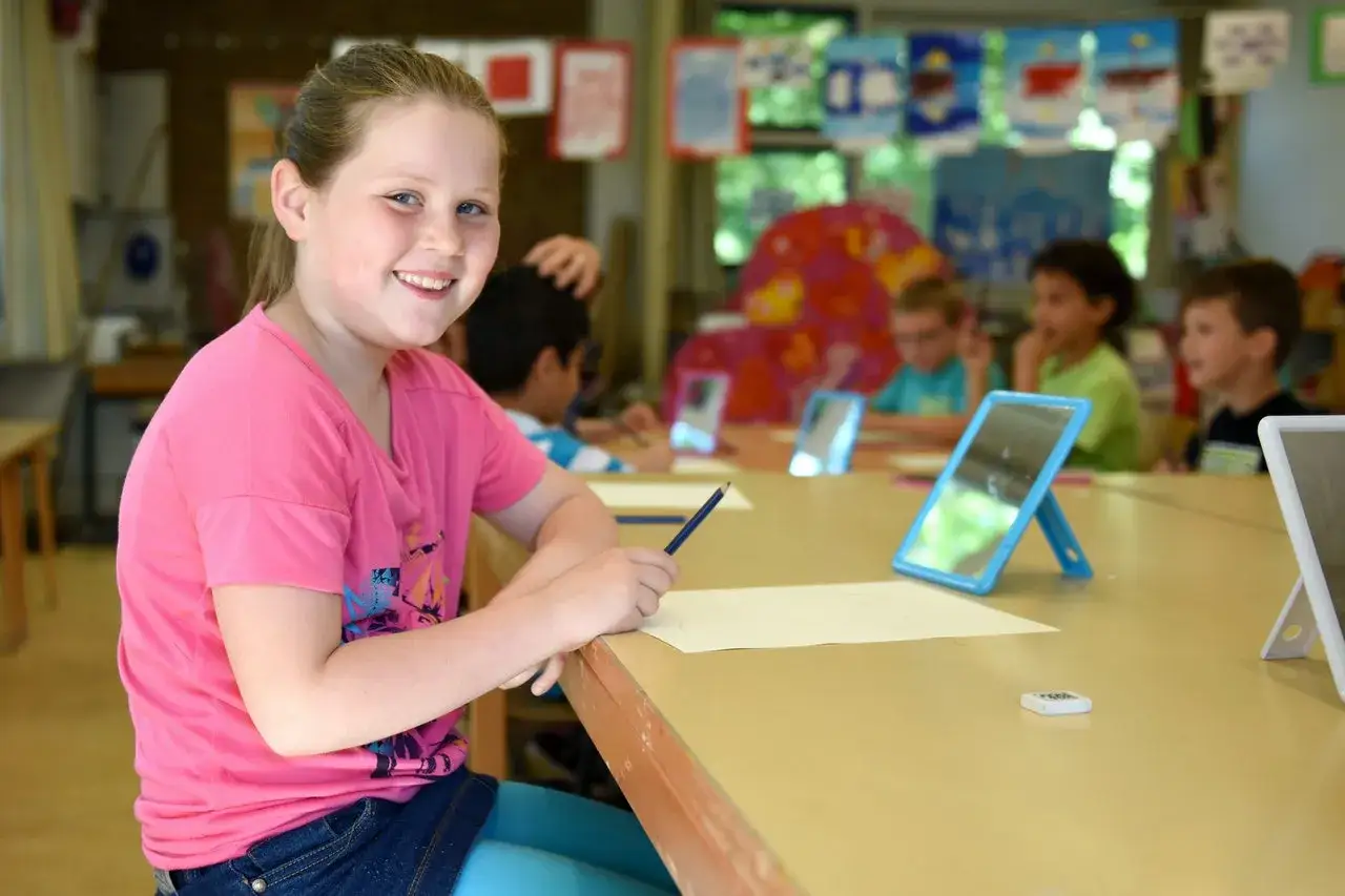 Een lachend meisje met een potlood in haar hand, omringd door klasgenoten. Dit is een voorbeeld van didactisch coachen in de klas.