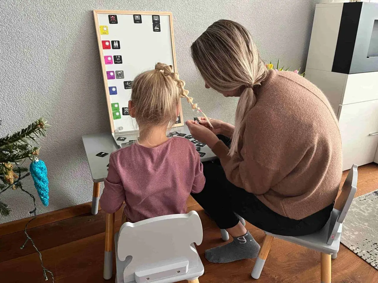 Een vrouw helpt een meisje met het vlechten van haar haar, terwijl ze samen op het spectrum zitten en naar een bord met symbolen kijken.