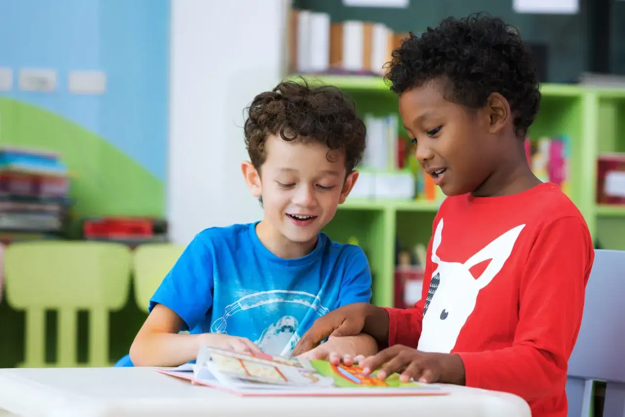 Twee lachende kinderen, een met een blauw shirt en een met een rood shirt, lezen samen een boek.
