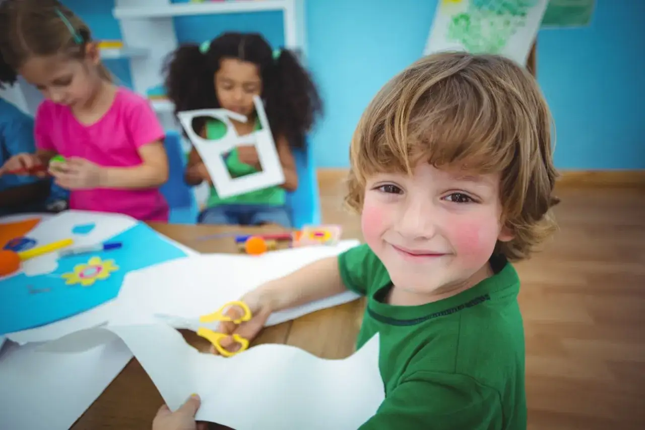Jongetje met schaar knipt papier, lachend. Andere kinderen knutselen ook. Dit kan helpen bij onderprikkeling autisme.