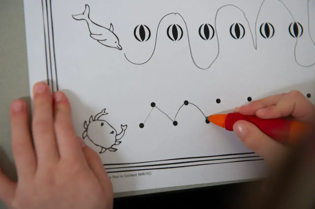 Kind oefent met een stift op een werkblad voor leren schrijven groep 1. Een visje volgt een lijn.