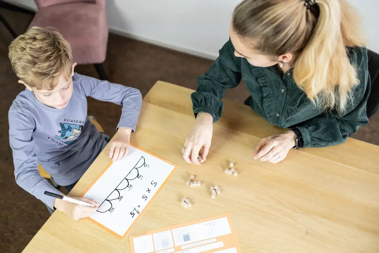Een jongen leert wat redactiesommen zijn met blokjes en een whiteboard. Hij schrijft 5 x 5 = 25.