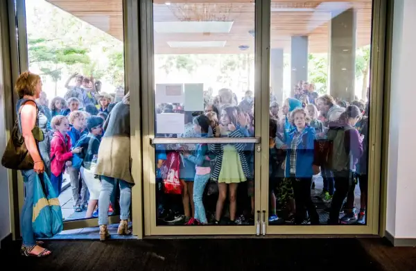 Kinderen staan te dringen bij een glazen deur, nieuwsgierig naar wat er binnen gebeurt. Ze lijken te wachten op iets, misschien wel de uitleg over wat zijn redactiesommen.