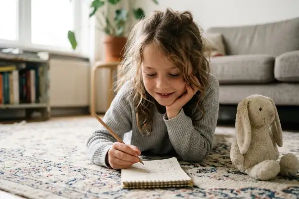 Een meisje ligt op de grond en maakt sommen in een schrift. Naast haar ligt een knuffelkonijn. Ze oefent de tafels.