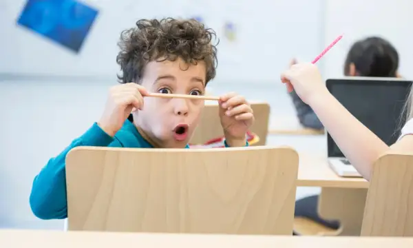 Een jongen van 6 jaar met krullend haar houdt een potlood voor zijn ogen, vol concentratie. Een ander kind steekt een roze potlood omhoog.
