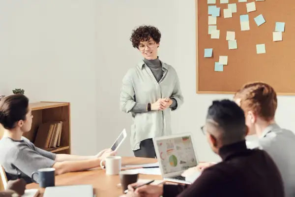 Een vrouw presenteert ideeën voor spreekbeurt onderwerpen aan een team. Post-its op het prikbord suggereren brainstormsessies.