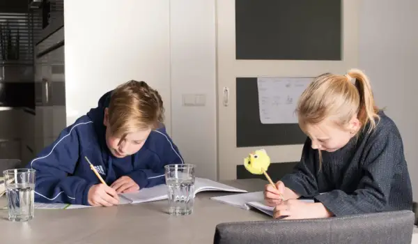 Twee kinderen maken huiswerk aan tafel. De jongen schrijft met een potlood, de meid met een leuke gele pen. Ze oefenen met delen met kommagetallen.